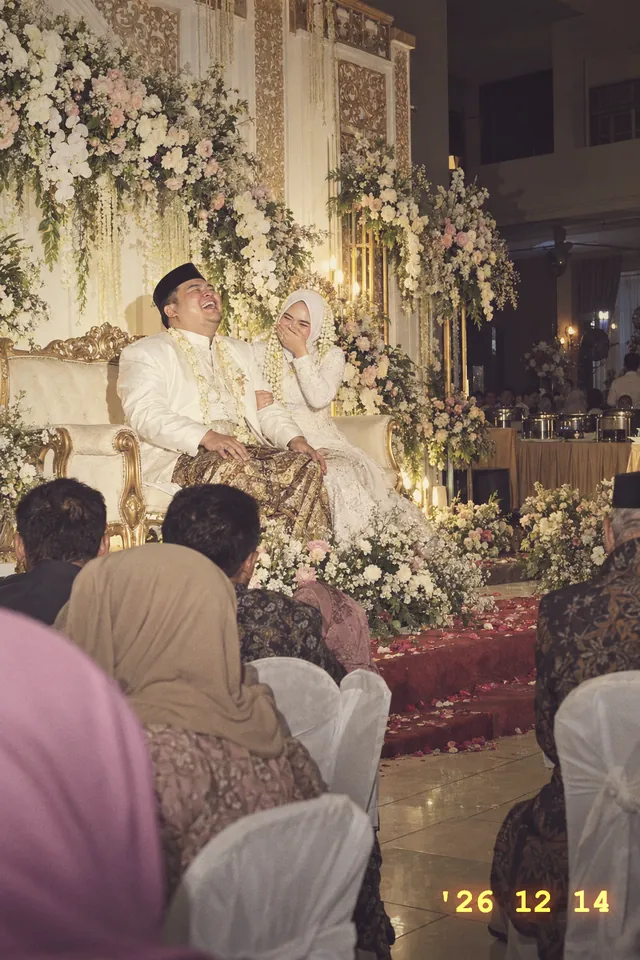 Nadia dan Raka tertawa lepas di pelaminan bunga putih, difoto dengan lampu kilat